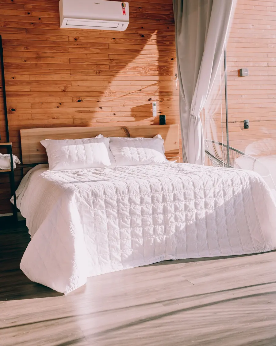 Cama de casal com colcha branca num chalé de madeira com cortinas leves
