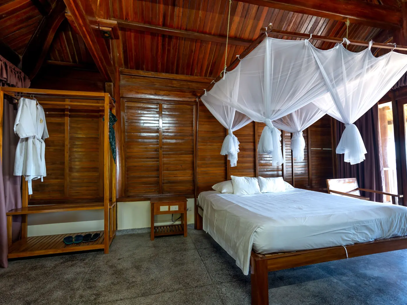 Suíte de madeira com cama de casal, mosquiteiro branco e porta de vidro para a varanda